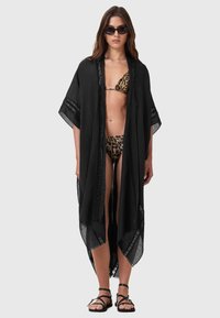 Copertura leggera in stile kimono nera con dettagli in pizzo, maniche ampie e apertura frontale. Indossata sopra un bikini con stampa leopardata.