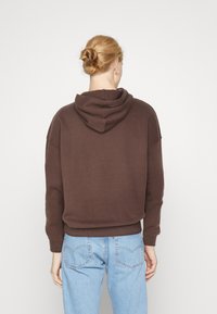 Hnedý krátky hoodie s kapucňou, so žebrovanými manžetami a pásom, v kombinácii so svetlo modrými džínami, zobrazený zozadu.