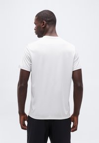 Άνδρας με την πλάτη γυρισμένη, φοράει μια απλή λευκή κοντομάνικη μπλούζα και μαύρο παντελόνι, μπροστά σε ανοιχτό γκρι φόντο.