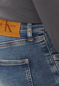 Jeans de denim en azul medio con un parche de cuero, costuras visibles y un bolsillo trasero. La tela parece suave con un ligero desgaste.