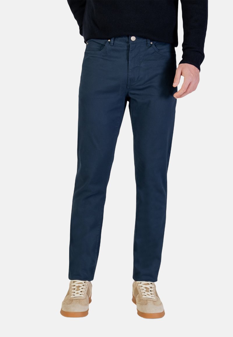 Pantaloni di cotone blu navy con taglio dritto, dotati di chiusura con bottoni, cinque tasche e una texture liscia, abbinati a sneakers beige.