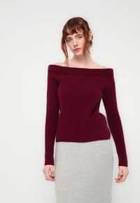 Μπορντό πουλόβερ με ανοιχτούς ώμους σε απαλό, υφασμάτινο πλέκτου, συνδυασμένο με μια ανοιχτό γκρι εφαρμοστή φούστα. Οι απλές, καθαρές γραμμές ενισχύουν τη σιλουέτα.