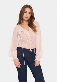 Blusa rosa trasparente con scollatura arricciata e maniche lunghe, dotata di dettaglio regolabile e polsini gather, abbinata a jeans blu scuro.