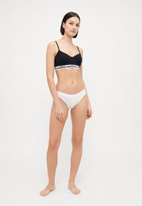 Crna bralette s pojasom s logom i bijelim donjim gaćicama niskog struka. Tkanina je glatka s podesivim trakama i minimalnim šavovima.