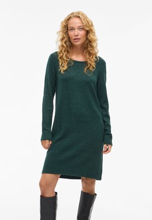 Robe en tricot à manches longues de couleur vert foncé avec un col rond, coupe décontractée, manches et ourlet à bords côtelés. Portée avec des bottes noires jusqu'aux genoux.