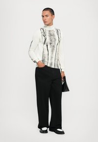 Μακριά μανίκια τούρτα σε off-white με μαύρο αφηρημένο σχέδιο· συνδυασμένο με φαρδιά μαύρα παντελόνια και μαύρες αξεσουάρ.