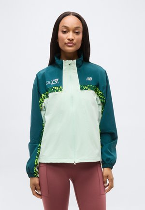 Mujer con chaqueta New Balance con cremallera en tonos verde agua y verde menta con detalles estampados en verde, y leggings color malva, posando contra un fondo blanco.