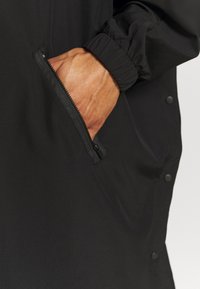 Schwarze Jacke mit einer Reißverschlusstasche, elastischen Bündchen und Druckknöpfen an der Seite. Der Stoff wirkt glatt und leicht.