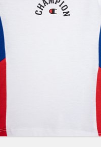 Bela majica s kratkimi rokavi z rdečimi in modrimi detajli, ki ima velik logotip "Champion" v črni barvi nad majhnim logotipom v rdeči in črni barvi.