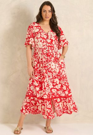 Kvinde stående iført en rød og hvid blomsterprintet midi-kjole med knapper, flæseærmer og sandaler, foran en neutral væg.