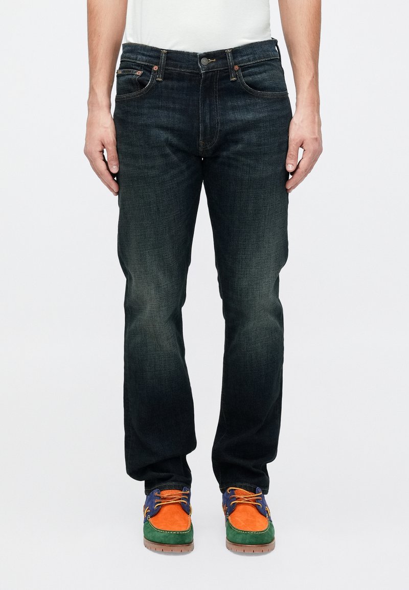 Hombre vestido con jeans oscuros y zapatos de ante coloridos con paneles en naranja, verde y azul, de pie contra un fondo blanco.