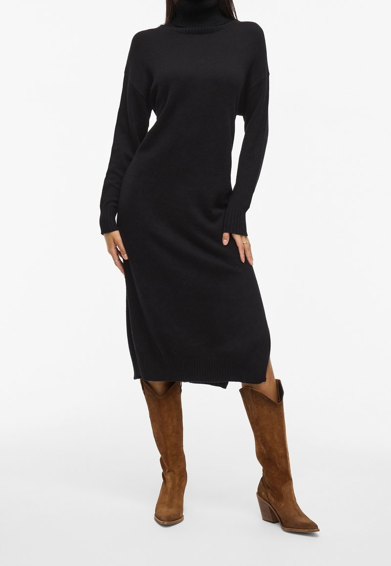 Femme portant une robe tricotée noire à manches longues et col roulé avec des fentes latérales, ainsi que des bottes de cowboy en daim marron mi-mollet, posant les mains près des hanches.