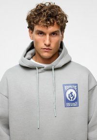 Lichtgrijze hoodie met een voorzak, capuchon met trekkoord en een blauwe grafische label met de tekst "PERSPECTIVE." Zachte textuur, ontspannen pasvorm.