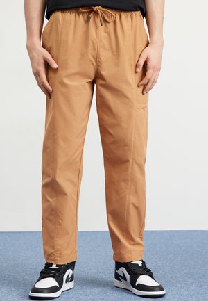 Beige Elastikbundhosen mit Kordelzug, Seitentaschen und einer Cargotasche, kombiniert mit schwarz-weißen Sneakern.