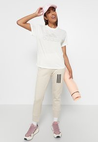 T-shirt bianca in cotone con logo "adidas", pantaloni della tuta beige con strisce nere, scarpe da ginnastica rosa e cappellino rosa chiaro, mentre tiene un tappetino da yoga.