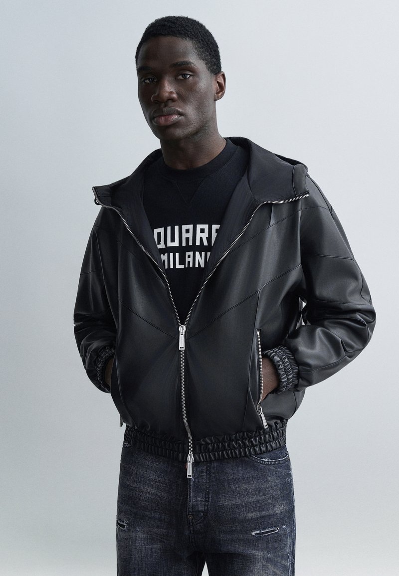 Fekete bőrből készült bombázó dzseki cipzáros előlappal, elasztikus mandzsettákkal és szegéllyel. Fekete grafikás pulóver és kopott sötét farmernadrág fölött viselve.