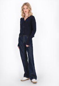 Cardigan tricotat în nuanță de bleumarin, cu decolteu în V și nasturi, asortat cu blugi largi din denim închisi la culoare și pantofi albi cu talpă bej.