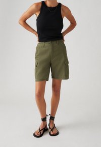 Vihreät cargo-shortsit, joissa etu- ja sivutaskut, yhdistettynä mustaan hihattomaan paitaan. Malli käyttää mustia remmisandaaleja. Sileä kangaspinta.