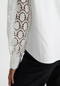 White blouse with sheer lace sleeves featuring an oval pattern, smooth fabric, and buttoned cuffs. Pairing with black pants.