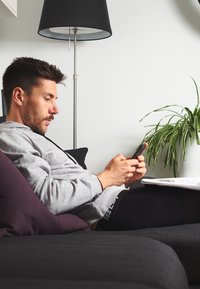Person in a grey hoodie using a black smartphone while seated on a dark sofa. A black floor lamp and green plant are in the background.