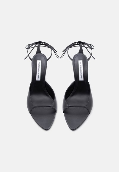 Ζευγάρι μαύρες γόβες με λεπτή μύτη και λουράκια που δένουν στον αστράγαλο, με ετικέτες "Victoria Beckham" στις εσωτερικές σόλες, σε λευκό φόντο.