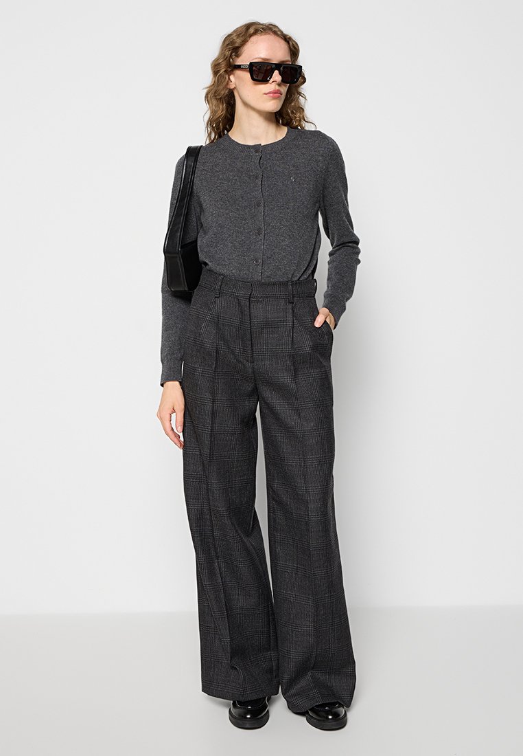 Grijze gebreide cardigan gecombineerd met hoog getailleerde, geruite broeken. Het model draagt grote zwarte zonnebril en heeft een zwarte tas.