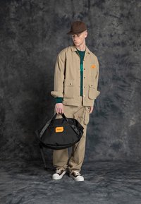 Beige work jacket with two front pockets, paired with matching pants, holding a black duffel bag featuring an orange logo. Brown cap and sneakers.