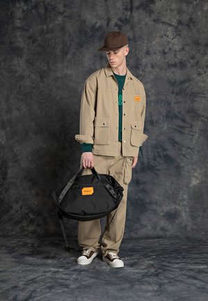 Beige work jacket with two front pockets, paired with matching pants, holding a black duffel bag featuring an orange logo. Brown cap and sneakers.