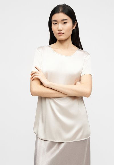 Νεαρή γυναίκα με μακριά σκούρα μαλλιά, ντυμένη με κρεμ σατέν μπλούζα με κοντά μανίκια και φούστα που ταιριάζει, στέκεται με σταυρωμένα τα χέρια μπροστά σε απλό φόντο.