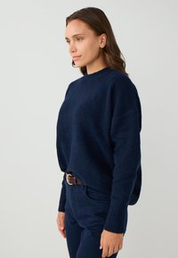 Maglione a maglia blu navy con finitura testurizzata, colletto a girocollo e polsini a coste, abbinato a pantaloni blu scuro e una cintura marrone.