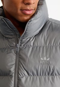 Grå pufferjakke med høj krave, frontlynlås og vandret syninger. Har et broderet Adidas-logo på brystet.