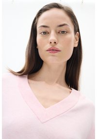 Gaiši rozā v-veida kakla džemperis ar ribinātu kakla daļu, mīkstu tekstūru un brīvu piegriezumu. Gluda auduma fons uz neitrāla fona.