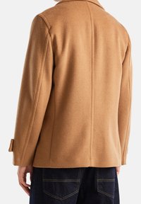 Abrigo marrón de mezcla de lana con cuello de muesca, corte recto y detalles de bolsillos de ojal, combinado con unos vaqueros de mezclilla oscura.