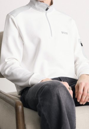 Person trägt einen weißen Pullover mit Viertelreißverschluss, grauem Kragen und dem "BOSS"-Logo, sitzt mit den Händen auf dunkler Hose.