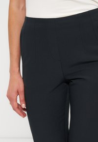 Personne portant un pantalon noir taille haute et un haut blanc, le bras gauche détendu le long du corps, vue de la taille à mi-cuisse.
