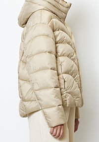 Béžová péřová bunda s kapucí, s prošívaným vzorem, hladkou texturou a zapínáním na zip. Rukávy jsou mírně přestřižené pro větší pohodlí.