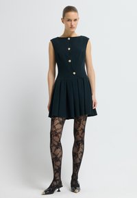 Robes sans manches vert foncé avec une jupe évasée et cinq boutons dorés. Le mannequin porte des collants noirs à motifs en dentelle et des chaussures noires à bout pointu.