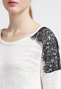 Femme portant un pull en tricot blanc avec un détail en dentelle noire sur l'épaule, lèvres visibles, fond neutre.