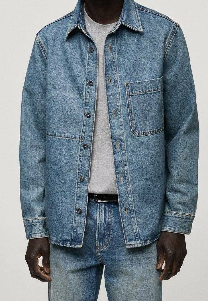 Lichtblauwe spijkerjack met een knoopsluiting aan de voorkant, een borstzak en lange mouwen. Draagt over een grijs t-shirt en bijpassende jeans.