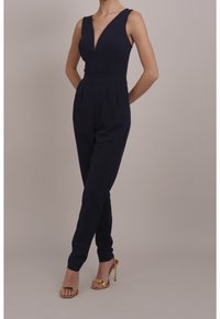 Mono de marina con escote en V, ajuste entallado y piernas ajustadas. Presenta una textura suave y tirantes anchos, combinado con tacones dorados.