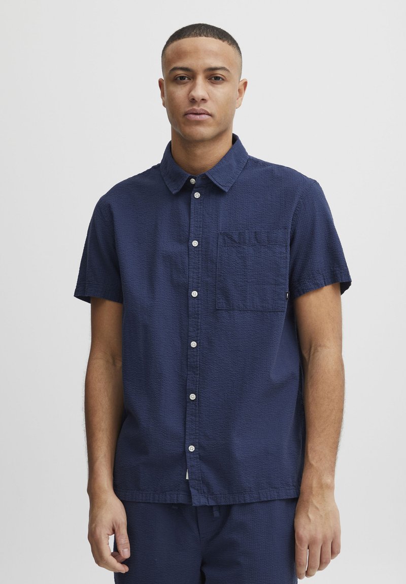 Camisa de manga corta en tejido texturizado azul marino oscuro con cierre de botones en el frente, cuello puntiagudo y un bolsillo en el pecho.