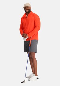 Helder oranje lange mouwen pullover met een rits, grijze shorts, witte sneakers en een beige pet, terwijl hij een golfclub vasthoudt met een witte handschoen.