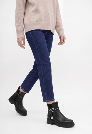 Personne portant un jean bleu, un pull beige et des bottines noires avec un détail boucle, marchant sur un fond blanc uni.