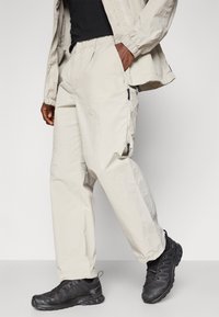 Light beige cargo pants with an elastic waistband and side pockets, paired with black athletic shoes featuring textured detailing.