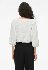Hvid tekstureret bluse med pufærmer og vid halsudskæring. Sammen med sorte højttaljede bukser, hvilket skaber et lagdelt look.