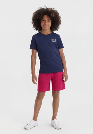 Dieťa s kučeravými vlasmi stojí oblečené v námornícky modrom tričku, červených šortkách a bielych teniskách na jednoduchom svetlom pozadí.