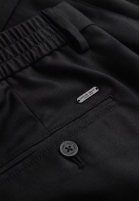 Gros plan sur un pantalon noir montrant une ceinture élastique, une poche arrière boutonnée et une petite étiquette métallique indiquant "ombre dept. since 1985."