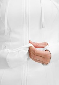Veste blanche zippée à texture lisse, avec des trous pour les pouces dans les poignets et un devant zippé. Des mains saisissent le tissu.