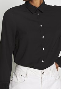 Chemise noire à manches longues avec col à revers, boutons blancs et poignets plissés, associée à un jean blanc taille haute.