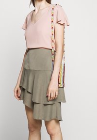 Jasnoróżowa bluzka z krótkim rękawem i dekoltem w serek, zestawiona z rozkloszowaną oliwkową spódnicą. Na ramieniu wisi mała, kolorowa torebka na ramię z koralików.